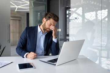 Stressed businessman with headache working on laptop in modern office, feeling overwhelmed and anxious