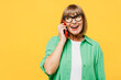 Elderly blonde woman 50s year old wear green shirt glasses casual clothes talk speak on mobile cell phone conducting pleasant conversation isolated on plain yellow background studio Lifestyle concept