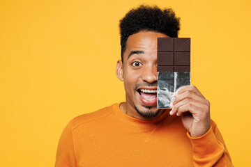 Wall Mural - Young surprised happy man wear orange sweatshirt casual clothes cover face with bar of chocolate isolated on plain yellow background studio Proper nutrition healthy fast food unhealthy choice concept