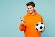 © ViDi Studio - Young happy man fan wears orange hoody casual clothes use mobile cell phone cheer up support football sport team hold in hand soccer ball watch tv live stream isolated on plain blue color background.
