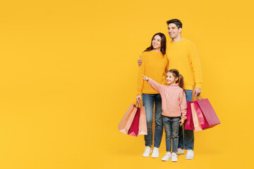 Wall Mural - Full body young parent mom dad with child kid girl 7-8 years old wear pink sweater casual clothes hold shopping bags point aside isolated on plain yellow background. Black Friday sale buy day concept.