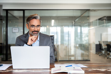 Mature Indian or Latin business man ceo trader using computer, typing, working in modern office, doing online data market analysis, thinking planning tech strategy looking at laptop with copy space.