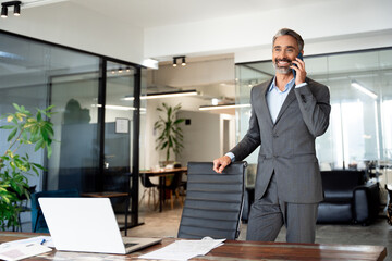 Wall Mural - Middle aged Latin or Indian businessman having call on smartphone with business partners or clients. Smiling mature Hispanic man in relaxing pose talking by mobile cellphone at work in modern office. 