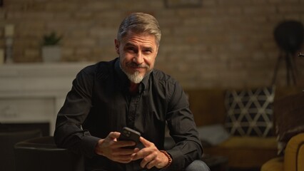 Wall Mural - Mid adult businessman with phone in home office, banking online, remote working. Portrait of happy older gray hair bearded man smiling. Entrepreneur managing business on internet.