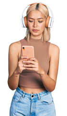 Sticker - Beautiful young blonde woman using smartphone wearing headphones relaxed with serious expression on face. simple and natural looking at the camera.