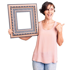 Poster - Beautiful young woman with short hair holding empty frame pointing thumb up to the side smiling happy with open mouth