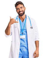 Wall Mural - Young hispanic man wearing doctor uniform and stethoscope smiling doing phone gesture with hand and fingers like talking on the telephone. communicating concepts.