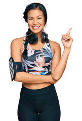 Wall Mural - Beautiful hispanic woman wearing sportswear and headphones with a big smile on face, pointing with hand and finger to the side looking at the camera.