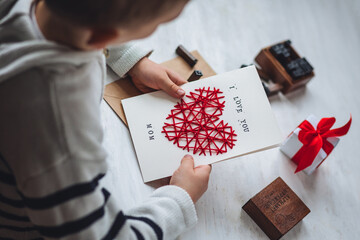 Concept of children craft, handmade gift for Mother's Day. Little boy holding craft card with embroidering in the shape of heart and printed message I love you Mom. Kid's leisure activity, handcraft