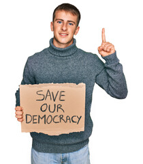 Poster - Young blond man holding save our democracy protest banner surprised with an idea or question pointing finger with happy face, number one