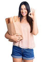 Poster - Young beautiful caucasian woman holding paper bag with bread doing ok sign with fingers, smiling friendly gesturing excellent symbol