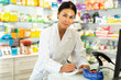 Asian female pharmacist in lab coat standing beside counter in drugstore and writing.