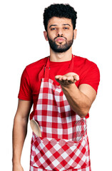 Wall Mural - Young arab man with beard wearing cook apron looking at the camera blowing a kiss with hand on air being lovely and sexy. love expression.