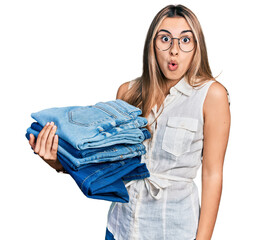 Canvas Print - Hispanic young woman holding stack of folded jeans scared and amazed with open mouth for surprise, disbelief face