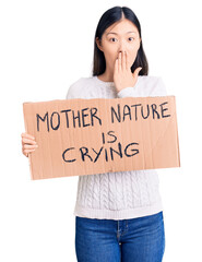 Wall Mural - Young beautiful chinese woman holding mother nature is crying cardboard banner covering mouth with hand, shocked and afraid for mistake. surprised expression