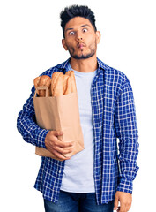 Wall Mural - Handsome latin american young man holding paper bag with bread making fish face with lips, crazy and comical gesture. funny expression.