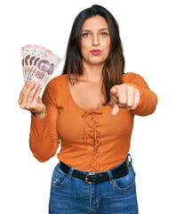 Wall Mural - Beautiful hispanic woman holding mexican pesos pointing with finger to the camera and to you, confident gesture looking serious