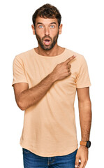 Sticker - Handsome young man with beard wearing casual tshirt surprised pointing with finger to the side, open mouth amazed expression.