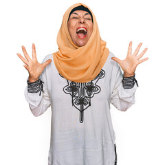 Poster - Middle age hispanic woman wearing traditional islamic hijab scarf crazy and mad shouting and yelling with aggressive expression and arms raised. frustration concept.