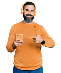 Wall Mural - Hispanic man with beard using smartphone wearing headphones smiling happy pointing with hand and finger