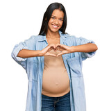 Fototapeta Panele - Beautiful hispanic woman expecting a baby showing pregnant belly smiling in love showing heart symbol and shape with hands. romantic concept.