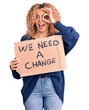 © Krakenimages.com - Young blonde woman with curly hair holding we need a change banner smiling happy doing ok sign with hand on eye looking through fingers