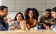 © Prostock-studio - Young diverse friends engaged in board game building tower indoors