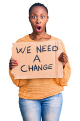 Sticker - Young african american woman holding we need a change banner scared and amazed with open mouth for surprise, disbelief face