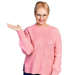 Wall Mural - Young blonde woman wearing casual clothes and glasses smiling cheerful presenting and pointing with palm of hand looking at the camera.