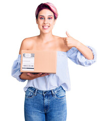 Sticker - Young beautiful woman with pink hair holding delivery package smiling happy and positive, thumb up doing excellent and approval sign