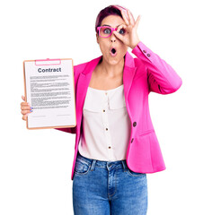 Poster - Young beautiful woman with pink hair holding clipboard with contract document doing ok gesture shocked with surprised face, eye looking through fingers. unbelieving expression.