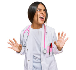 Wall Mural - Beautiful hispanic woman wearing doctor uniform and stethoscope crazy and mad shouting and yelling with aggressive expression and arms raised. frustration concept.