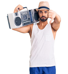 Sticker - Young hispanic man holding boombox, listening to music with angry face, negative sign showing dislike with thumbs down, rejection concept