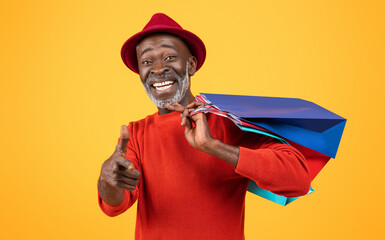 Wall Mural - Charismatic senior Black man with a white beard, giving a thumbs up and carrying colorful shopping bags