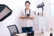 Portrait of young Asian photographer smiling while holding DSLR camera at photo and video production studio office.