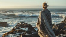  A Woman Standing On Top Of A Rocky Beach Next To The Ocean With A Blanket Draped Over Her And Looking At The Ocean.