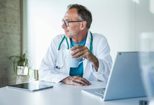 Contemplative Senior Doctor Sitting With Coffee Cup At Desk