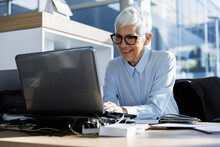 Smiling Senior Businesswoman Using Laptop Sitting At Desk In Office