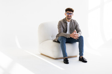 Wall Mural - Portrait of attractive man sitting in armchair and looking at camera isolated over white background