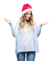 Canvas Print - Beautiful young blonde woman wearing christmas hat over isolated background clueless and confused expression with arms and hands raised. Doubt concept.