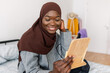 © Xavier Lorenzo - Happy african black woman looking at mirror while putting muslim headscarf at home