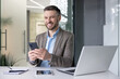 © Liubomir - Smiling businessman in a modern office using smartphone with laptop, notebook, and tablet on desk, conveying success and connectivity.