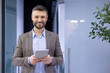 © Liubomir - Confident businessman in smart casual attire smiling, holding a tablet in modern office setting, embodying success and professionalism.