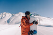 snowboarder holding snowboard and smartphone in his hands on prepared ski slope communicates by phone online and answers messages
