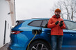 © Halfpoint - Man charging electric car during cold snowy day, using electric vehicle charging app, checking battery life, energy consumption on smart phone. Charging and driving electric vehicles during winter