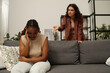 Young irritated brunette woman blaming her upset African American girlfriend sitting on couch and touching temple during quarrel