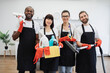 © sofiko14 - Portrait of happy multiethnic janitors holding cleaning equipment against background of light modern kitchen. Cleaning team, group of four multiracial people in black aprons and red gloves.