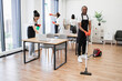 © sofiko14 - Young adult African American man wearing black apron and red rubber gloves vacuuming floor of modern light office. Professional service team of cleaning company.