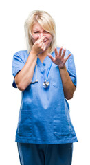 Wall Mural - Young beautiful blonde doctor woman wearing medical uniform over isolated background smelling something stinky and disgusting, intolerable smell, holding breath with fingers on nose. Bad smells