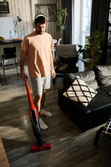 Wall Mural - Young man in casualwear listening to music in headphones and cleaning floor of living room with vacuum cleaner on weekend